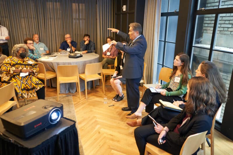 A speaker gestures as he shares his thoughts with a breakout session, surrounded by others seated and listening.